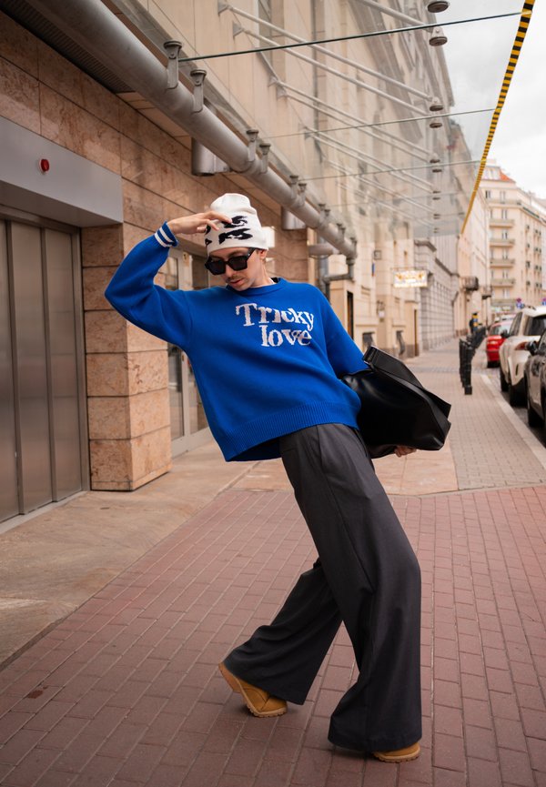 Persona con un suéter azul "Tricky love", pantalones negros anchos, gorro blanco, gafas de sol y sosteniendo una bolsa negra en una acera de la ciudad.