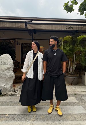 Un hombre y una mujer están fuera de un café, ambos llevan ropa negra con zapatos amarillos, junto a un letrero de piedra que dice "House of Wheat."