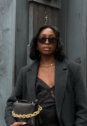 Top de satén negro con detalles de encaje, chaqueta gris ajustada, gafas de sol oversized negras y un bolso negro pequeño con cadena dorada.