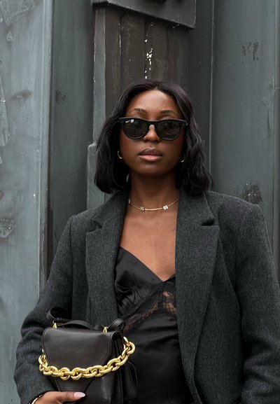 Top de satén negro con detalles de encaje, chaqueta gris ajustada, gafas de sol oversized negras y un bolso negro pequeño con cadena dorada.