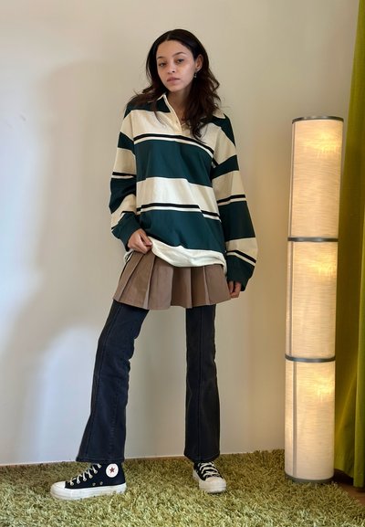 Top de rayas verdes y crema de manga larga, falda marrón con pliegues, jeans negros acampanados y zapatillas negras con suelas blancas. De pie sobre una alfombra verde.