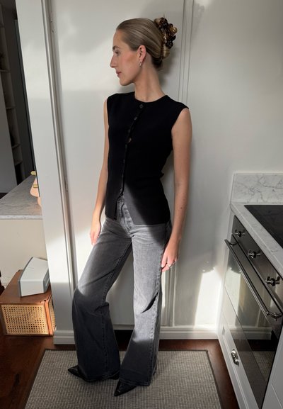 Top negro sin mangas con un cierre frontal de botones, combinado con jeans acampanados gris oscuro y tacones negros puntiagudos. Texturas suaves y diseño minimalista.