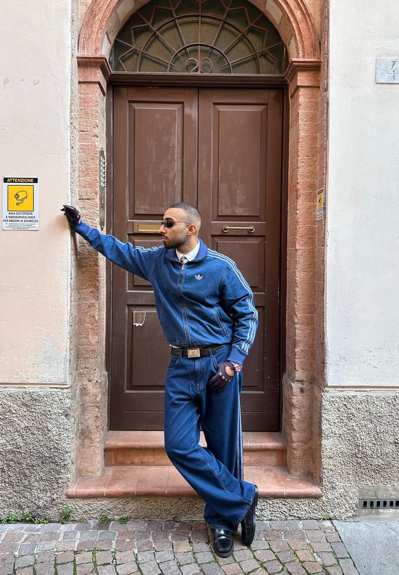 Άνδρας με μπλε φόρμα Adidas στηρίζεται σε τοίχο δίπλα σε κλειστή ξύλινη πόρτα με αψιδωτό παράθυρο πάνω από αυτήν, σε λιθόστρωτο δρόμο κάτω.