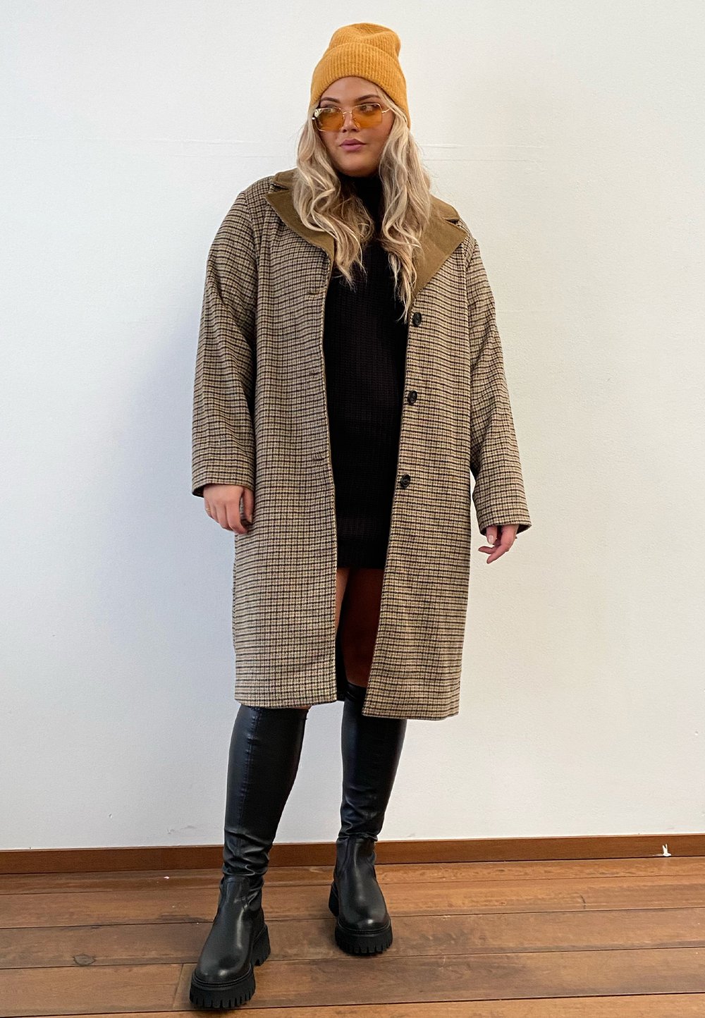 Casaco castanho em padrão pied-de-poule com grande gola e botões, combinado com um vestido preto e botas pretas até ao joelho. Gorro amarelo e óculos de sol incluídos.