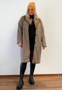 Casaco castanho em padrão pied-de-poule com grande gola e botões, combinado com um vestido preto e botas pretas até ao joelho. Gorro amarelo e óculos de sol incluídos.