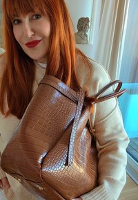 Femme aux cheveux roux portant un pull beige et tenant un grand sac à main marron à motif crocodile, à l'intérieur, près d'une fenêtre et d'une sculpture.