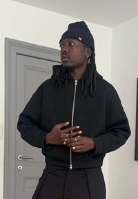 Sweat à capuche noir zippé avec une texture lisse et une fermeture éclair argentée ; porté avec un pantalon foncé et accessoirisé de bagues.