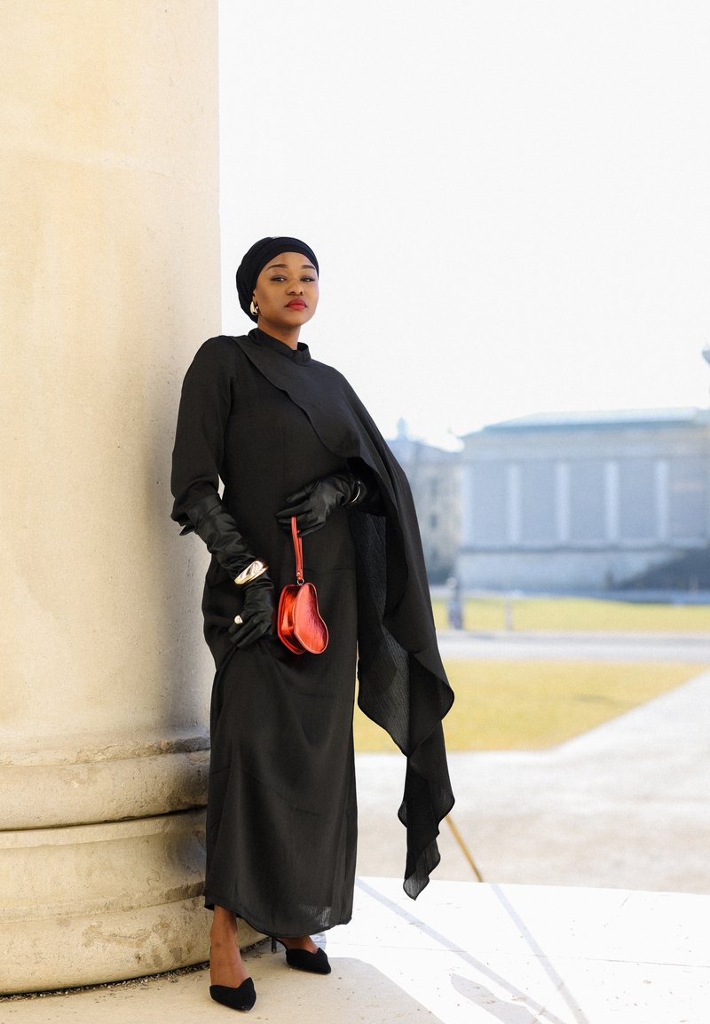 Femme en longue robe noire et foulard, debout près d'une grande colonne, portant des gants noirs et tenant un petit sac à main rouge en plein air.