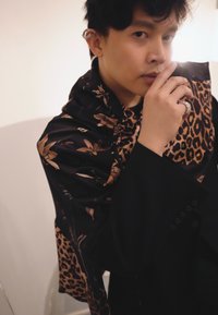 Veste noire avec écharpe imprimée léopard et motifs floraux, présentant une texture soyeuse et des motifs complexes. Bague au doigt, fond neutre.