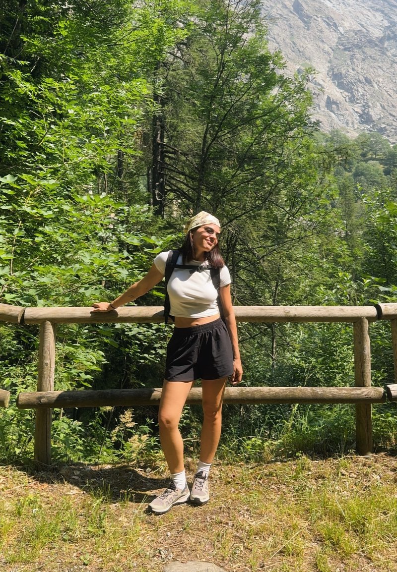 Jeune femme en tenue de randonnée appuyée contre une clôture en bois, entourée d'arbres verts et denses avec une montagne visible en arrière-plan.