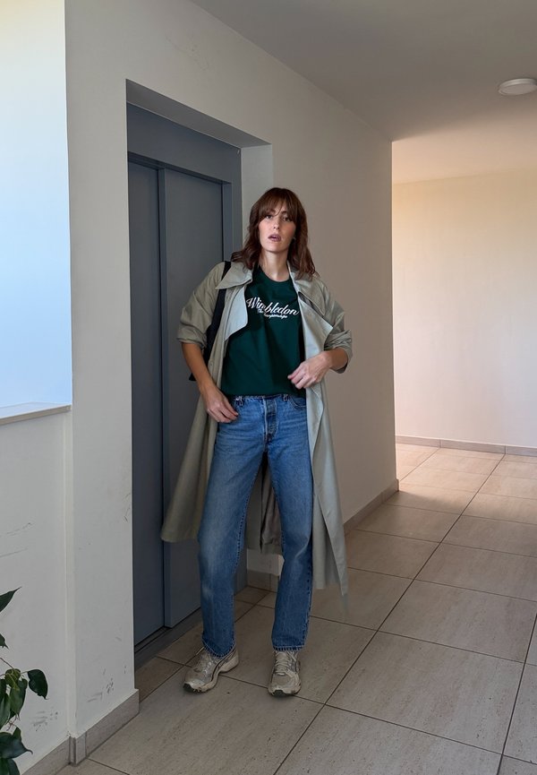 Una mujer posa en un pasillo junto a un ascensor oscuro. Lleva un abrigo largo beige, una camiseta verde, vaqueros azules y zapatillas deportivas.