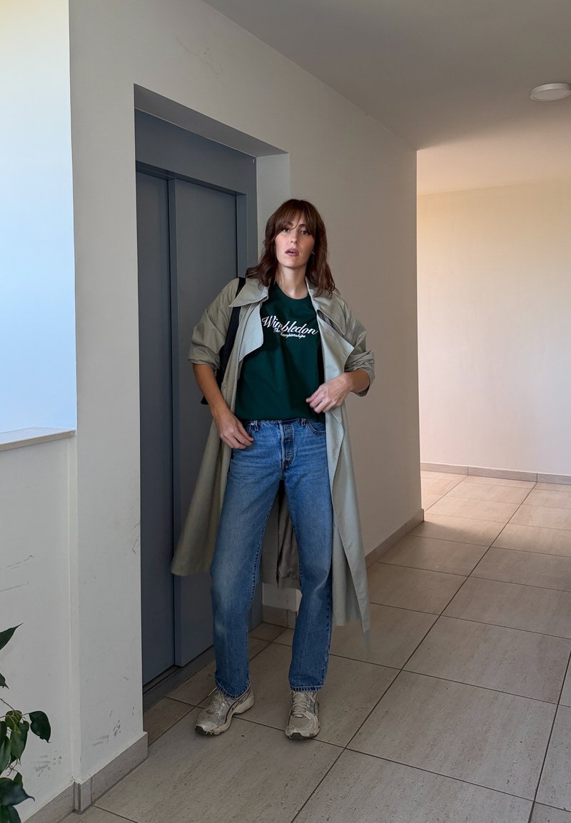 Une femme pose dans un couloir à côté d’un ascenseur sombre. Elle porte un long manteau beige, un T-shirt vert, un jean bleu et des baskets.