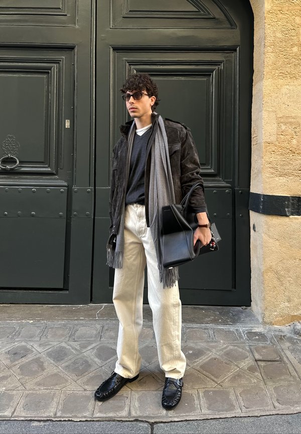 Mand iført mørk jakke, gråt halstørklæde, sorte solbriller, hvide bukser og sorte sko, der står ved store mørkegrønne døre og holder en sort taske.