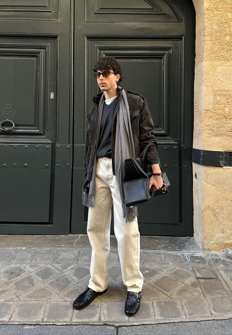 Homme portant une veste foncée, une écharpe grise, des lunettes de soleil noires, un pantalon blanc et des chaussures noires, debout près de grandes portes vert foncé en tenant un sac noir.