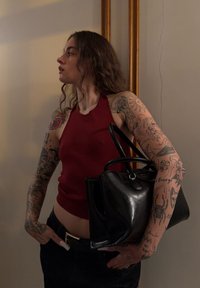 Jeune femme tatouée aux longs cheveux, portant un haut rouge sans manches et tenant un sac à main en cuir noir, debout à l'intérieur contre un mur beige.