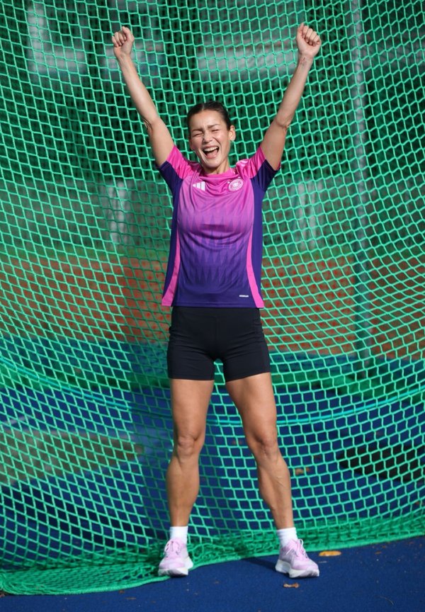 Atleta in una maglietta sportiva rosa e viola e pantaloni corti neri, che festeggia con le braccia alzate davanti a una rete verde in mesh.