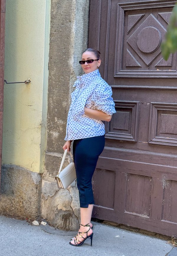 Femme portant des lunettes de soleil, une veste à pois, un pantalon noir et des talons hauts, debout à côté d'une grande porte en bois tenant un sac à main blanc.
