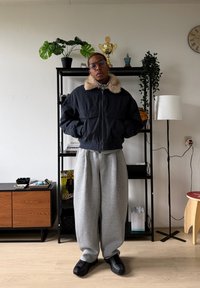 Veste bomber noire avec un col en fausse fourrure, pantalon de survêtement gris et baskets noires. En arrière-plan, des plantes en pot et une étagère.