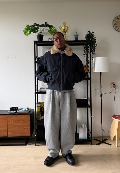 Chaqueta bomber negra con cuello de piel sintética, pantalones de chándal grises y zapatillas negras. El fondo presenta plantas en macetas y una estantería.