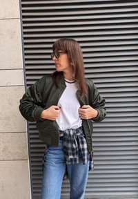 Blouson aviateur vert olive avec épaules texturées, porté sur un t-shirt blanc, associé à un jean bleu clair et une chemise à carreaux nouée.