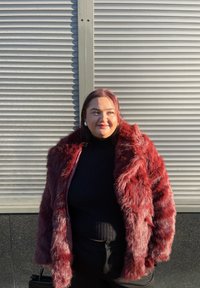 Manteau en fausse fourrure rouge avec manches longues, col large et apparence texturée, porté avec un col roulé noir et un pantalon noir.