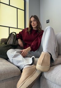 Beige ruskindstøfler med syningsdetaljer, grå joggingbukser og en bordeaux sweatshirt, siddende på en tekstureret beige sofa med en grøn taske.