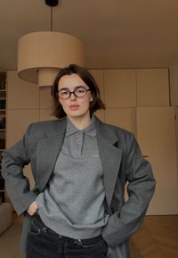 Blazer gris porté sur un polo gris, avec un col et un tissu texturé. Jean noir au design simple. Lumière intérieure douce.