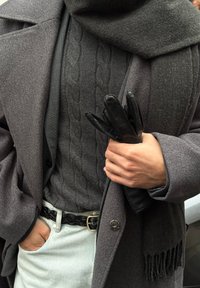 Siv volnen plašč s cofki, pod njim teksturiran siv pulover s kabelskim vzorcem, v roki držane črne usnjene rokavice in črnen pleten pas z okroglo sponko.