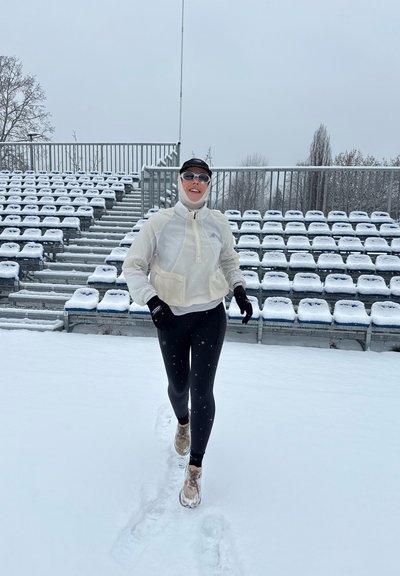 Persona con ropa de correr de invierno trotando sobre la nieve frente a asientos vacíos cubiertos de nieve en un estadio.