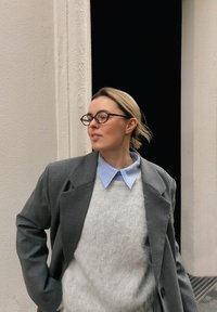 Grå oversized blazer over en hvit, lodden genser med en lyseblå skjorte med krage. Runde briller og en mørk bakgrunn med teksturert vegg.