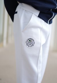 Pantaloni da tennis bianchi con finitura texturizzata, dotati di tasche laterali e di un patch del Lacoste Tennis Club con grafiche di racchette.