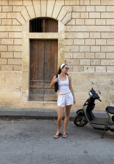 Joven mujer vestida con una camiseta blanca sin mangas y pantalones cortos, de pie junto a un scooter negro cerca de una pared de piedra texturizada con una puerta de madera.