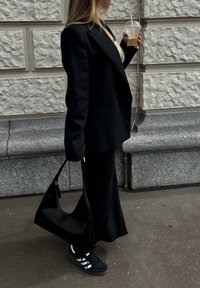 Crni oversized sako preko uske crne suknje, uparen s crnim tenisicama i crnom torbom. Osoba drži napitak u prozirnoj čaši.