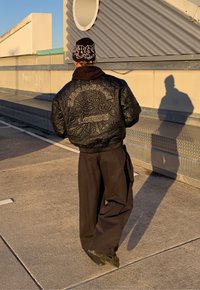 Jachetă bomber neagră matlasată cu broderia „SHANGHAI LORDS”, combinată cu pantaloni negri largi. Fundalul prezintă beton și o structură circulară.