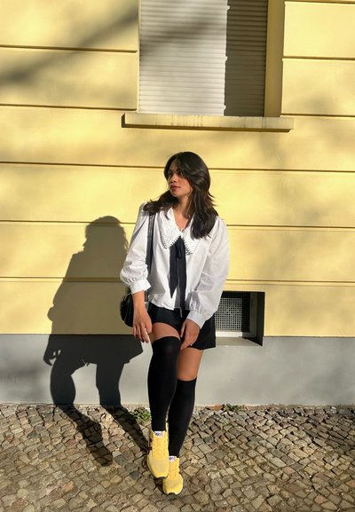Blusa blanca con un gran cuello, cinta negra, pantalones cortos negros y calcetas negras hasta la rodilla; zapatillas amarillas sobre pavimento de adoquines.