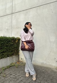Svetle šedé kostkované kalhoty, pruhovaná košile s knoflíky, burgundská kabelka, béžové tenisky; pozadí tvoří betonová zeď a zelený keř.