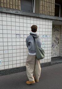 Person, die einen grauen Kapuzenpullover und beigefarbene weite Hosen trägt, mit einer grünen Umhängetasche, steht vor einer gefliesten Wand mit Graffiti.