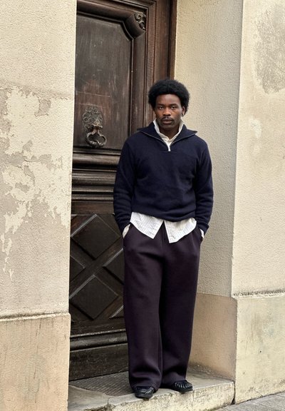 Hombre con peinado afro, vestido con suéter negro, camisa blanca y pantalones negros holgados, de pie contra una pared texturizada junto a una puerta de madera.