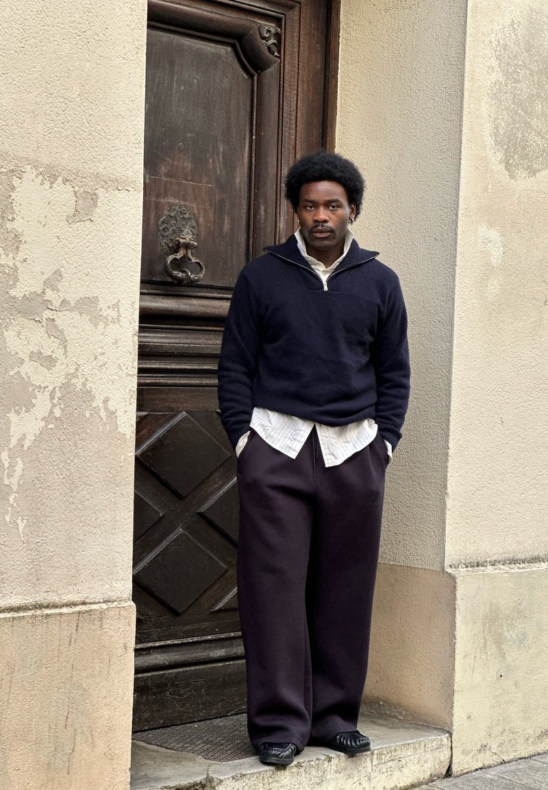 Homme avec coiffure afro portant un pull noir, une chemise blanche et un pantalon noir ample, debout contre un mur texturé près d'une porte en bois.