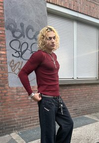 Top ajustat de culoare mahon cu mâneci lungi în dungi, asociat cu pantaloni negri largi cu buzunare și lanțuri. Perete texturat în fundal.