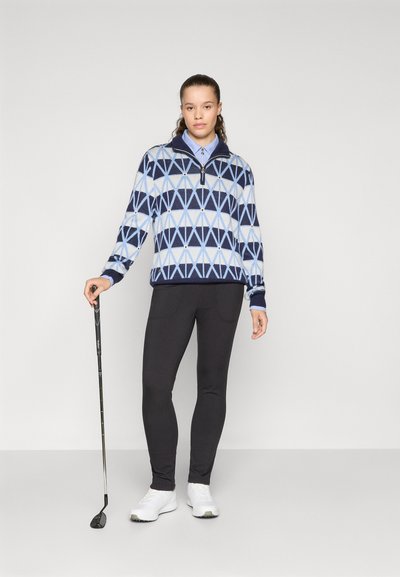 Pulóver de punto con un patrón geométrico en azul y marino, cuello con cremallera, leggings negros y zapatos de golf blancos, sosteniendo un palo de golf.
