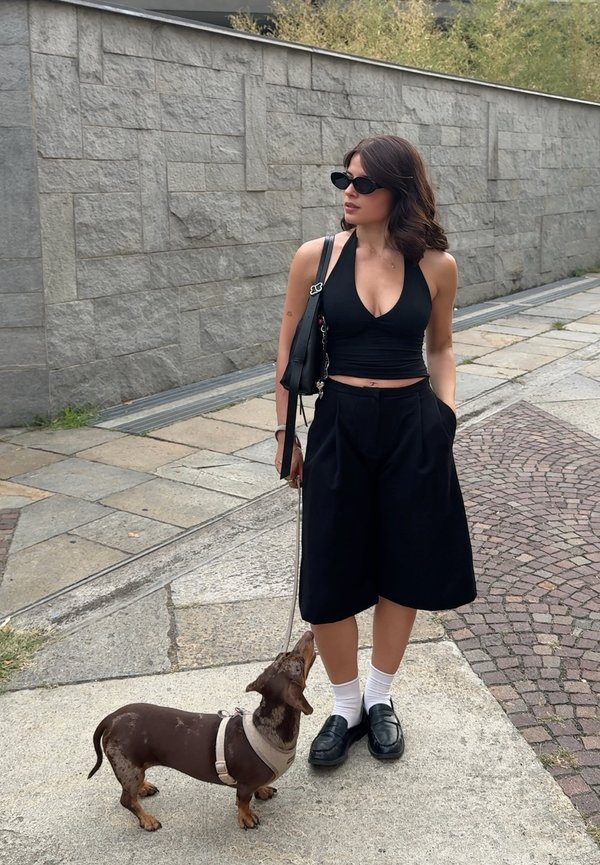 Vrouw in zwarte top en culottes staat op de stoep en kijkt weg, terwijl ze een bruine teckel aan de lijn naast zich vasthoudt.