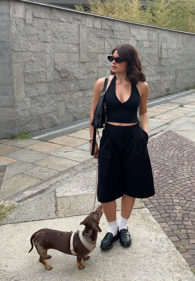 Femme en haut noir et culottes se tient sur le trottoir en regardant ailleurs, tenant un teckel marron en laisse à côté d'elle.