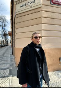 Mustmust värvi tekstuuriga mantel laia kraega, mille all on hall kampsun ja valge särk. Ringikujulised päikeseprillid ja must kott, seisev veepoe ees.