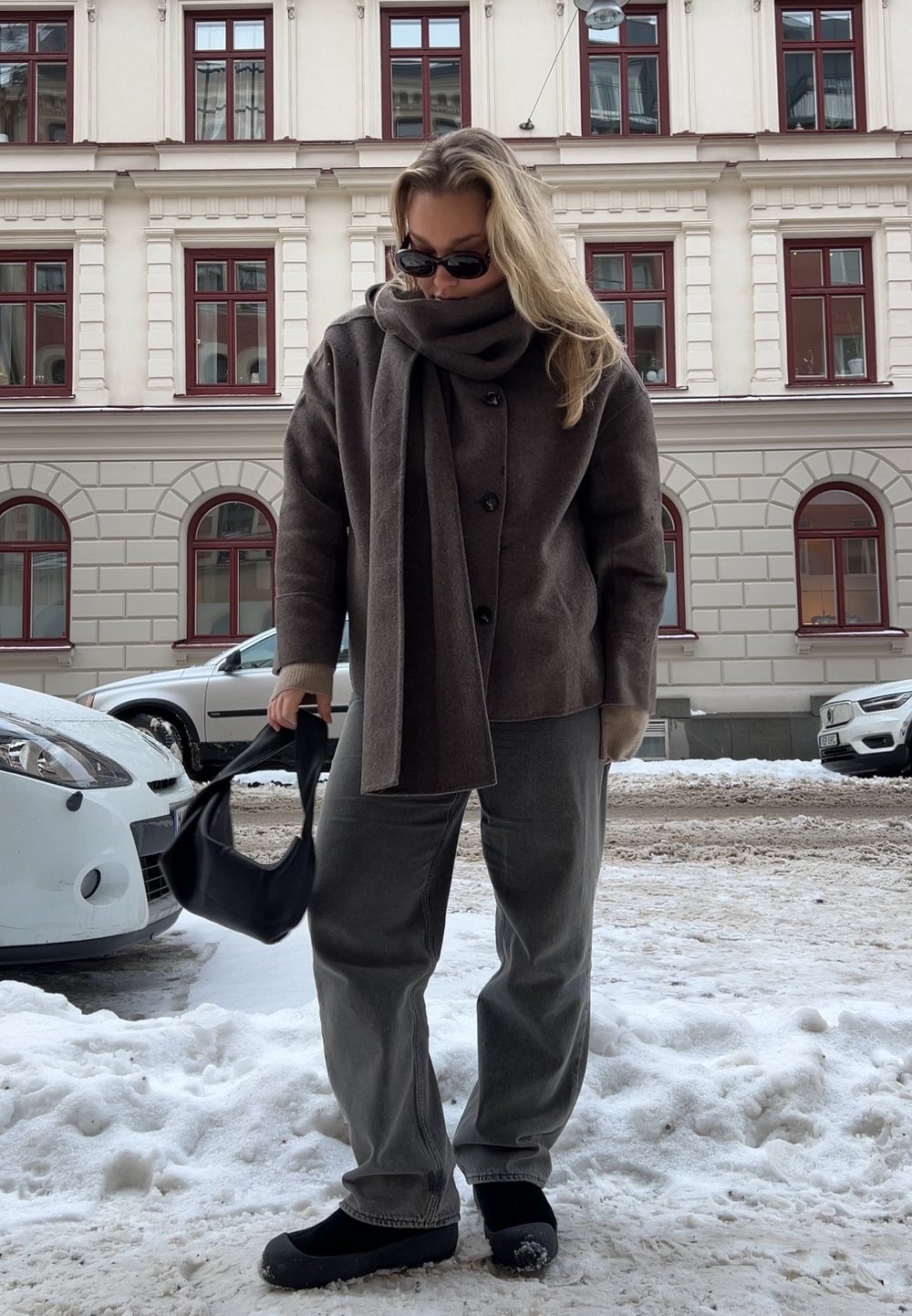 Ruda vilnos striukė su dideliais mygtukais, deranti šalikas, pilkos kelnės ir juodi batai, laikantis juodą rankinę, stovintis sniege.