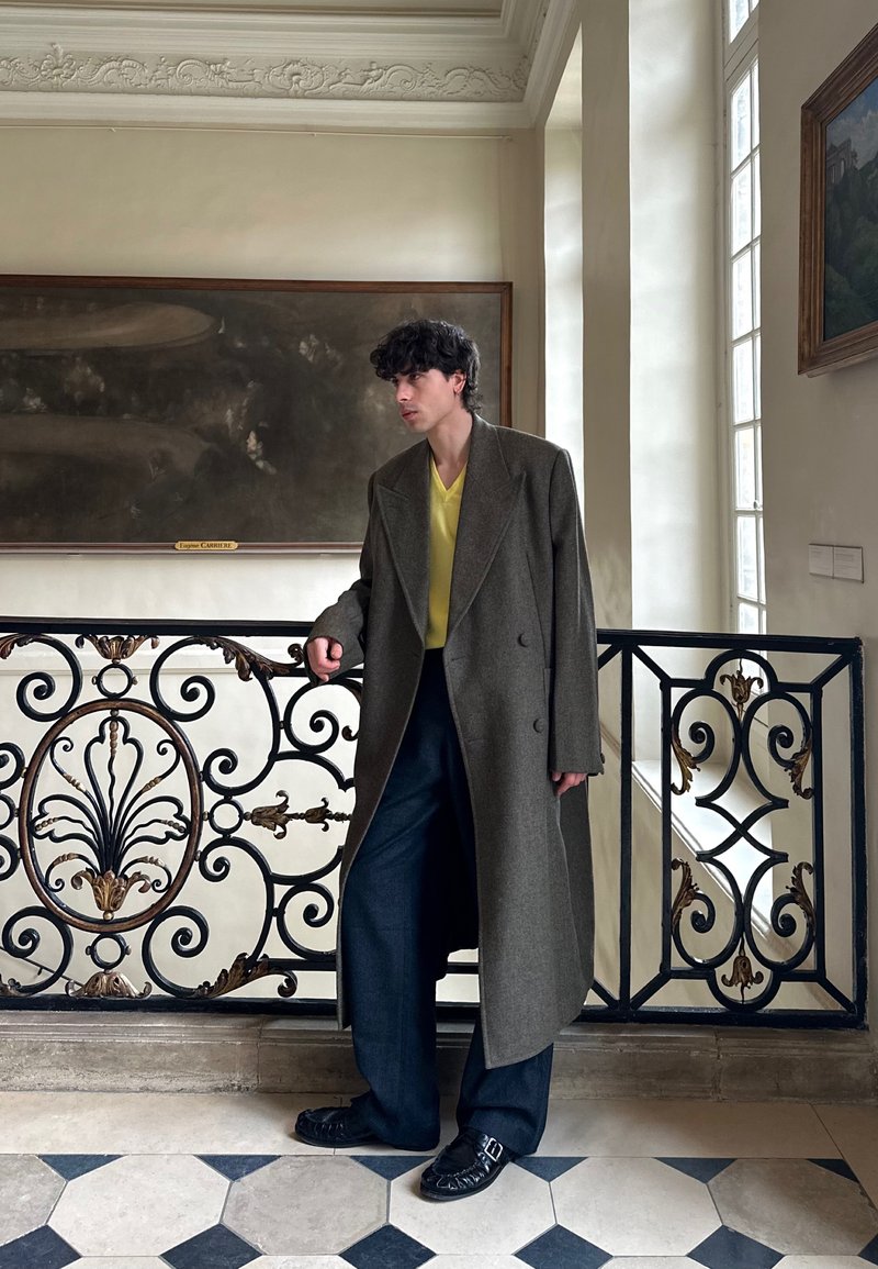 Man in long gray coat, yellow shirt, and dark pants leaning on ornate black and gold railing inside a bright room with paintings and tall windows.