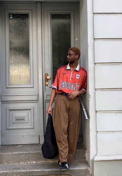 Jersey rojo y blanco "REFORMULAT", pantalones de pierna ancha marrones, zapatillas negras, sosteniendo un bolso negro, de pie junto a una puerta gris.