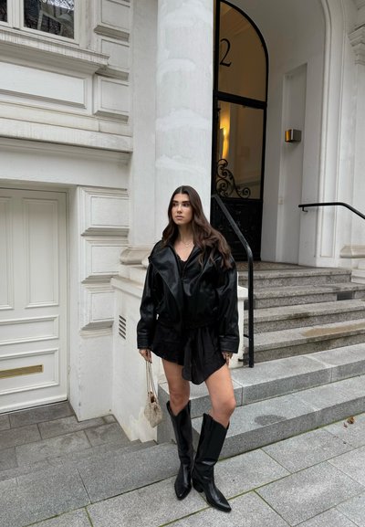 Chaqueta extragrande de cuero negro superpuesta sobre un top texturizado negro y falda. Botas negras hasta la rodilla. Escaleras y edificio blanco como fondo.