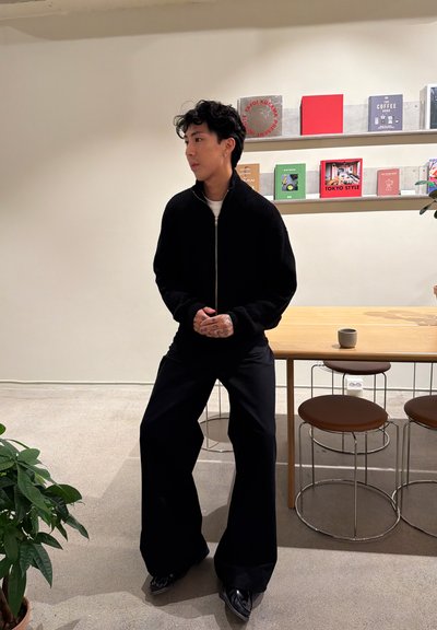 Joven vestido con suéter negro con cremallera y pantalones anchos negros, de pie junto a una mesa de madera con taburetes y estanterías en una habitación luminosa.