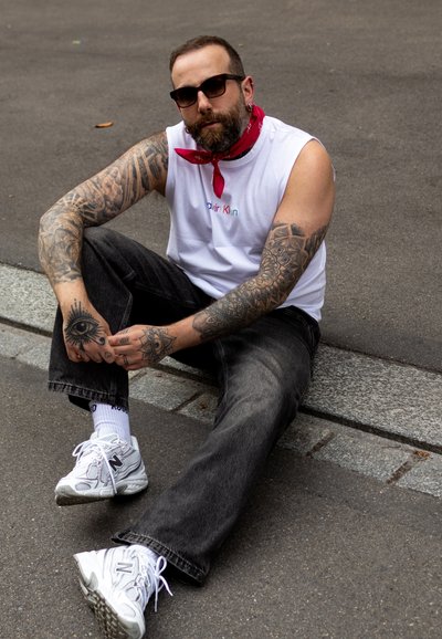 Camisa sin mangas blanca con el logo de "Calvin Klein", bandana roja, jeans negros, zapatillas blancas de New Balance y tatuajes detallados en los brazos.